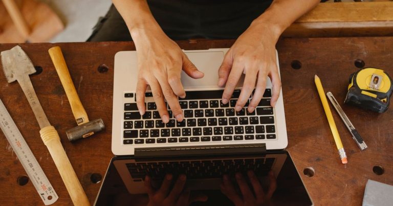 Mains d'un artisan tapant sur un ordinateur portable posé sur un établi avec ses outils